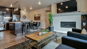 A modern kitchen with a central island, a dining area with chairs, a living room with a sofa, a flat-screen TV, and a fireplace. - Moab Arches C4 "Sage Creek" - Heated Pool-Hot Tubs (Moab)