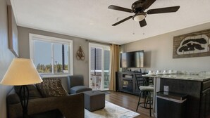 A modern living room with a sofa, a coffee table, a lamp, a ceiling fan, and a kitchen area with a TV and bar counter. - ML 323 - Modern, bike-in/bike-out summer retreat! (Snowshoe)