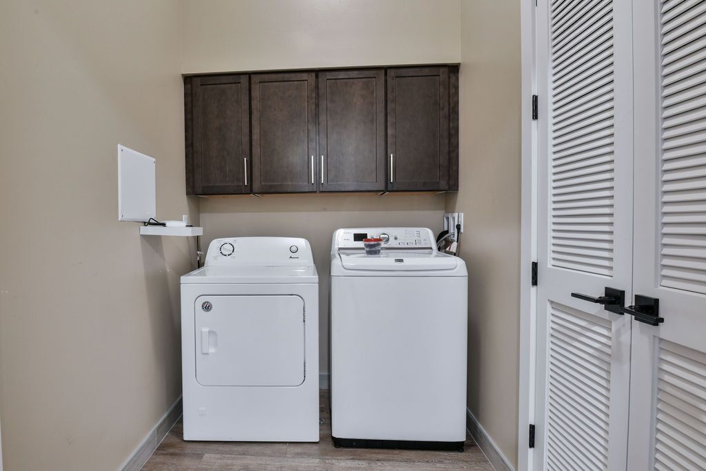 A laundry room with two white washers and dryers, dark wooden cabinets above, and a white door with louvered panels to the right.