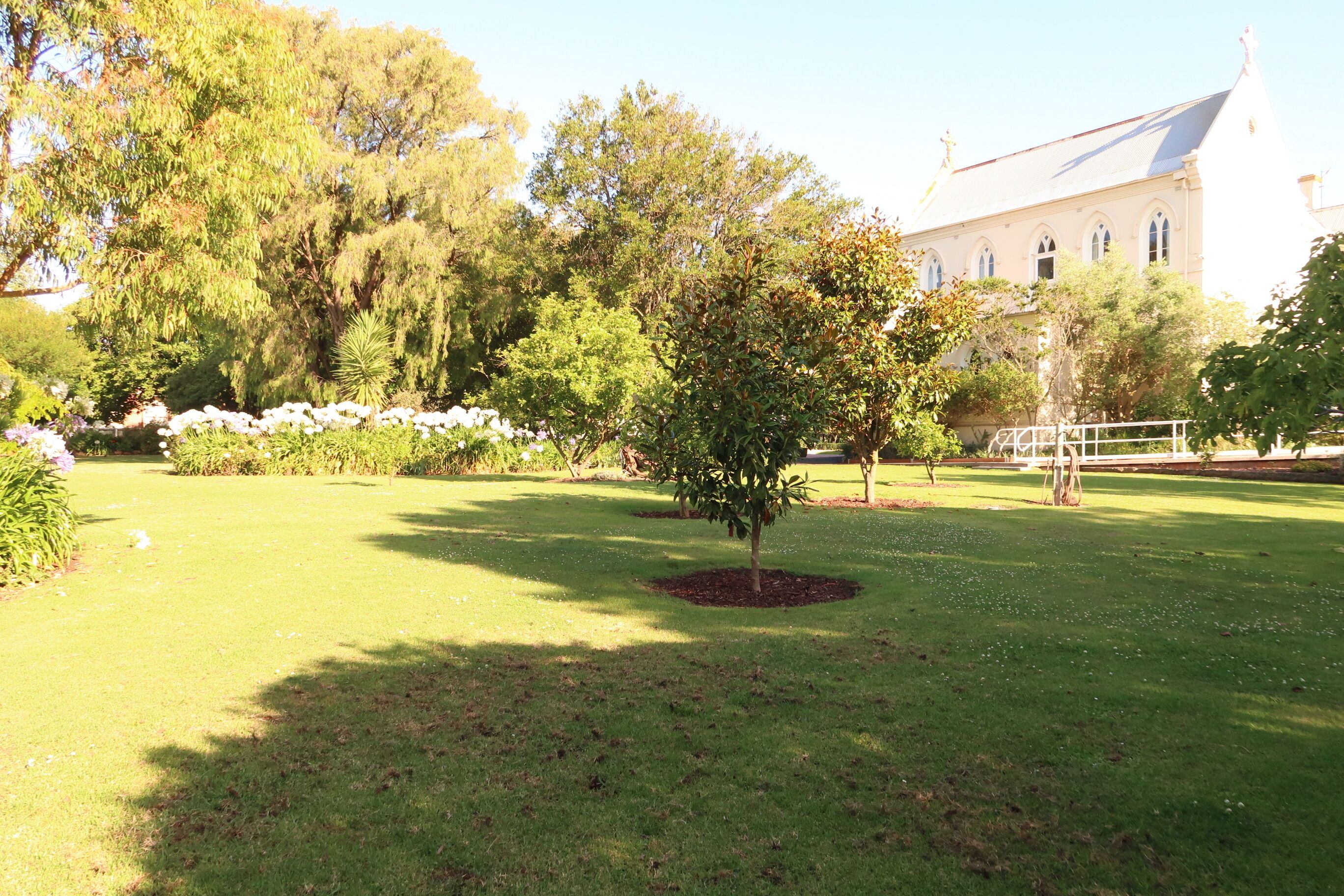 A well-maintained garden with a variety of trees and shrubs, a lawn, and a building with a pitched roof in the background.