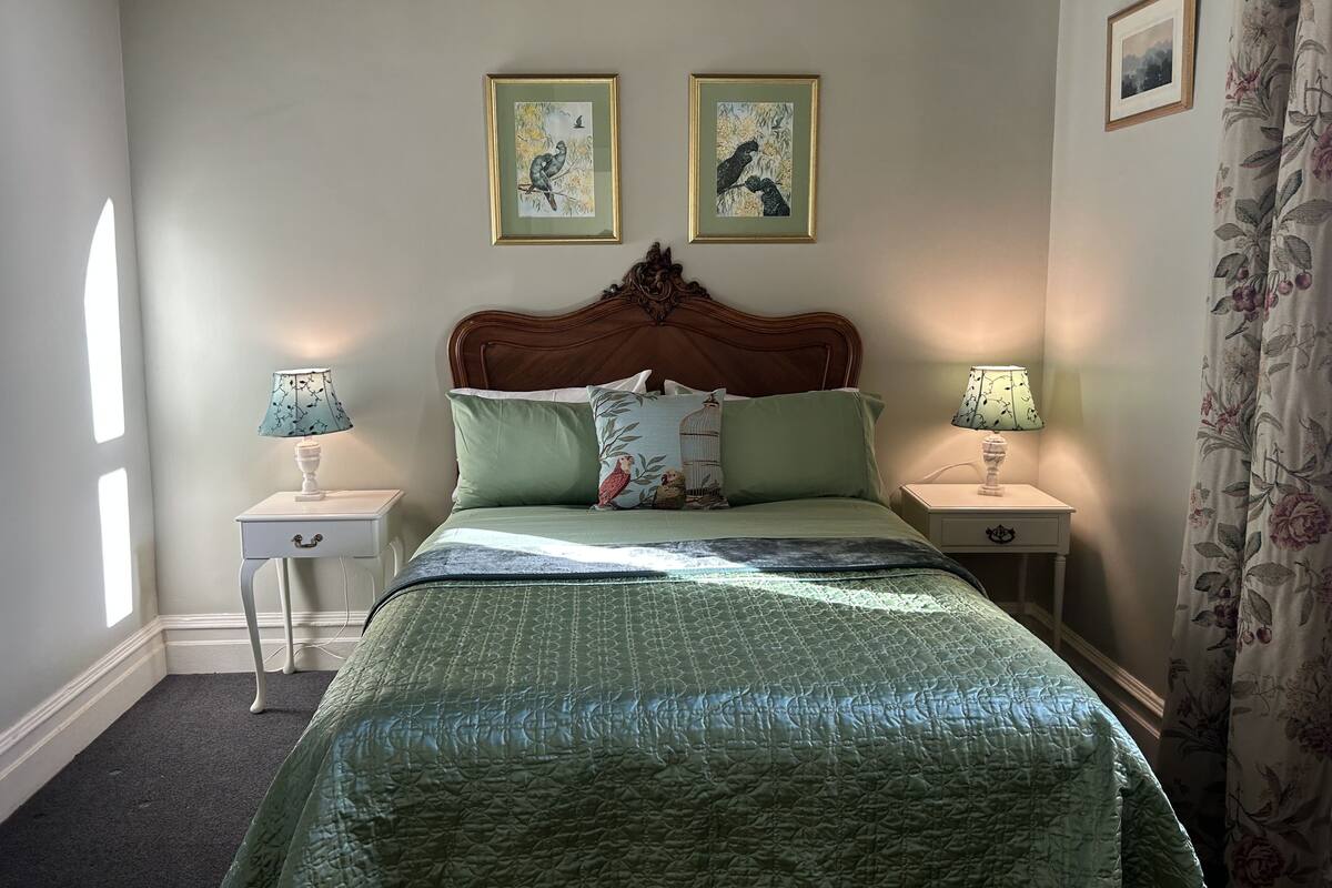 A neatly arranged bedroom with a wooden headboard, two bedside tables with lamps, a green quilted bedspread, and two framed pictures on the wall.