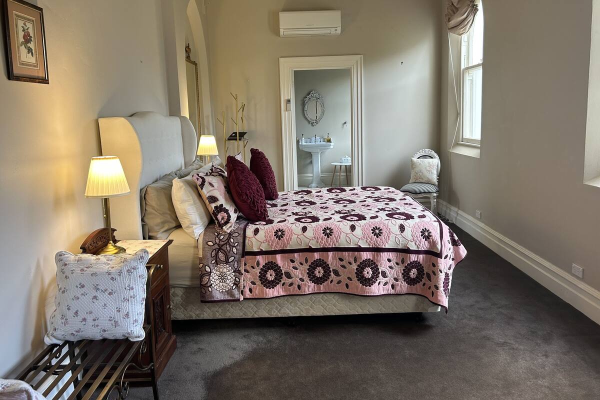 A bedroom with a bed, bedside table, lamp, mirror, and a window.