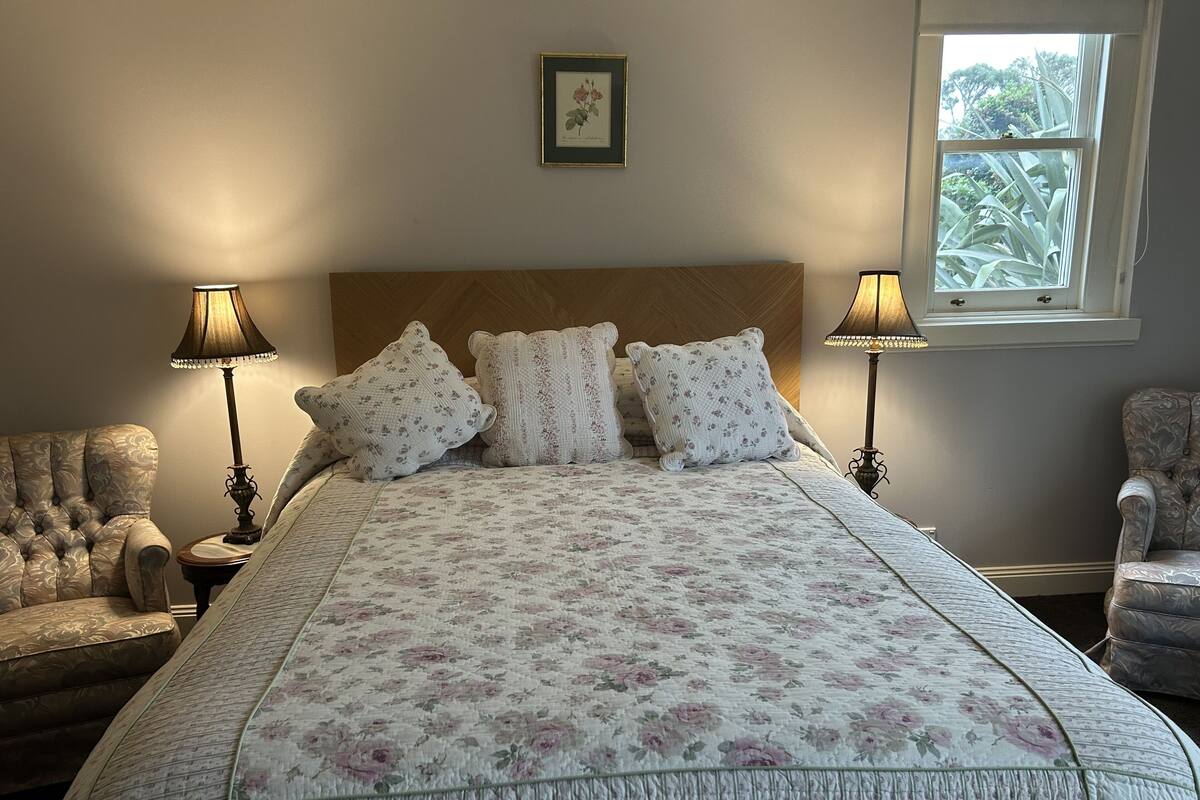 A bedroom with a large bed, floral bedding, two lamps, a chair, and a window with a view of trees.