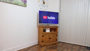 A room with a wooden cabinet under a flat-screen TV displaying the YouTube logo, a clock on the wall, and a window with blinds. - Captivating 2-bed Apartment in Edgbaston (Birmingham)