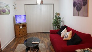 A living room with a red sofa, a glass coffee table, a TV on a wooden stand, a large window with blinds, and a plant. - Captivating 2-bed Apartment in Edgbaston (Birmingham)
