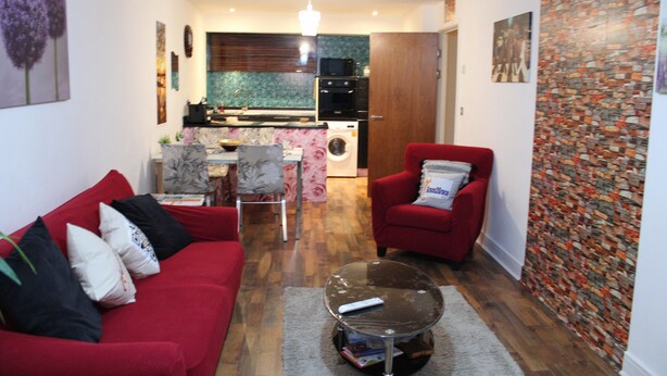 A compact living space with a red sofa, a glass coffee table, a red armchair, and a kitchen area with a washing machine. - Captivating 2-bed Apartment in Edgbaston (Birmingham)