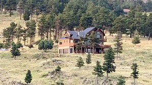 Ein zweistöckiges Haus mit großer Veranda und geneigtem Dach, umgeben von einem bewaldeten Gebiet.