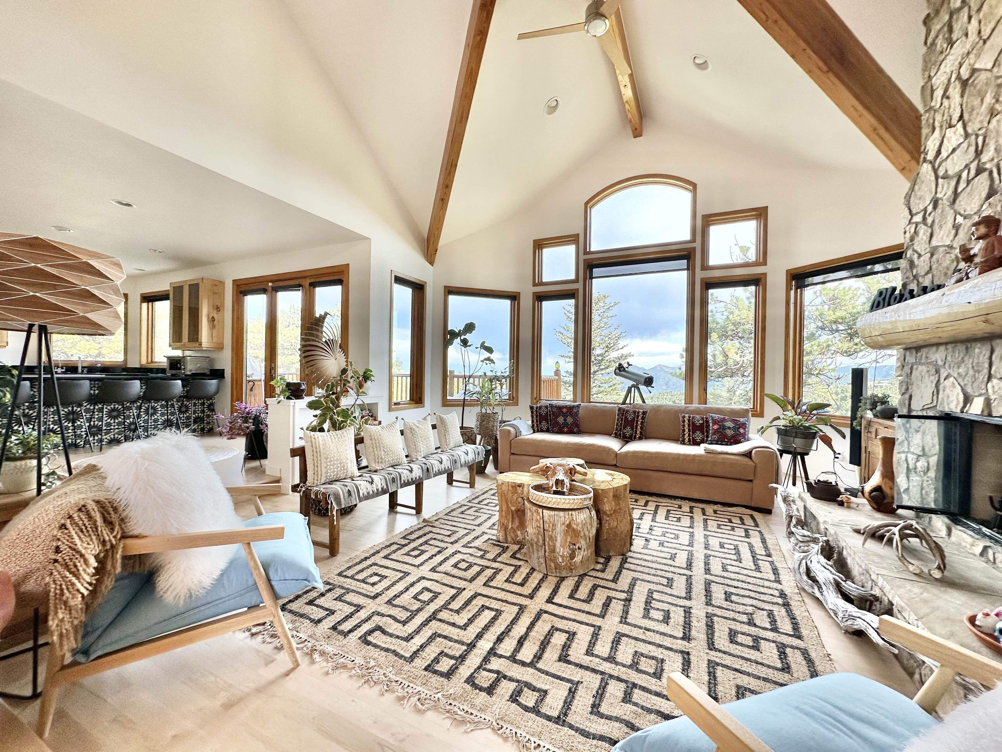 A spacious living room with a high ceiling, large windows, a stone fireplace, and a cozy seating area with a patterned rug.