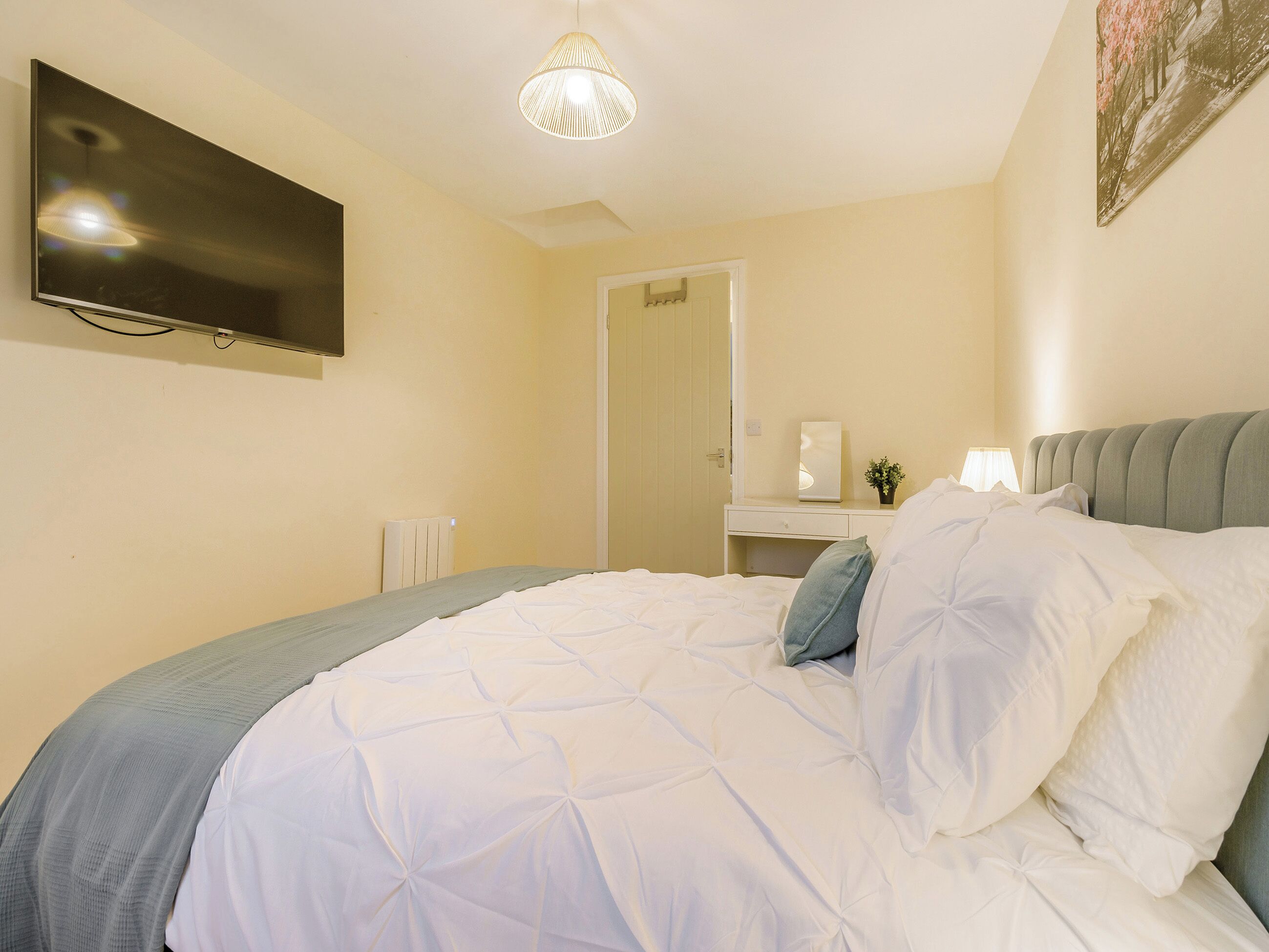 A neatly made bed with a grey headboard, a bedside table with a lamp, and a flat-screen TV mounted on the wall.