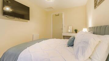 A neatly made bed with a grey headboard, a bedside table with a lamp, and a flat-screen TV mounted on the wall.