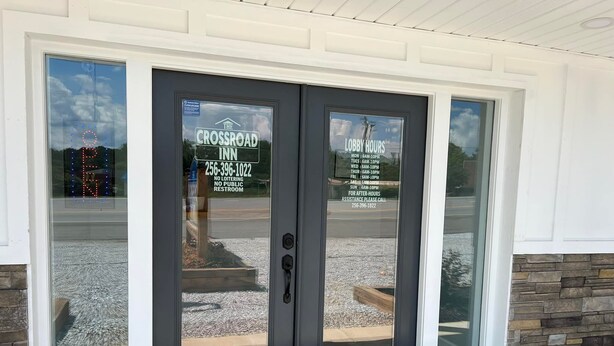 A double door entrance to The Crossroad Inn with a stone and wood exterior, a clear view of the parking lot, and a sign indicating lobby hours and a contact number for assistance. - The Crossroad Inn (Lineville)