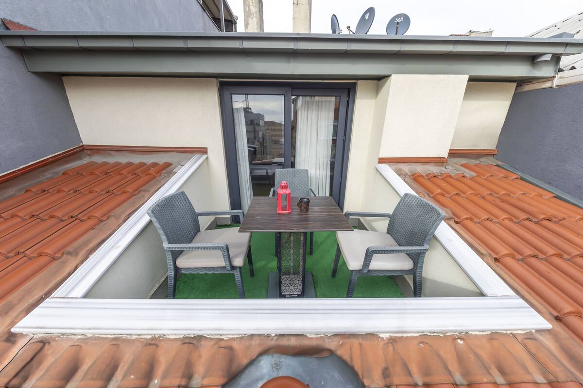 A rooftop terrace with a table and chairs, a small table with a red object, and a view of buildings in the background.