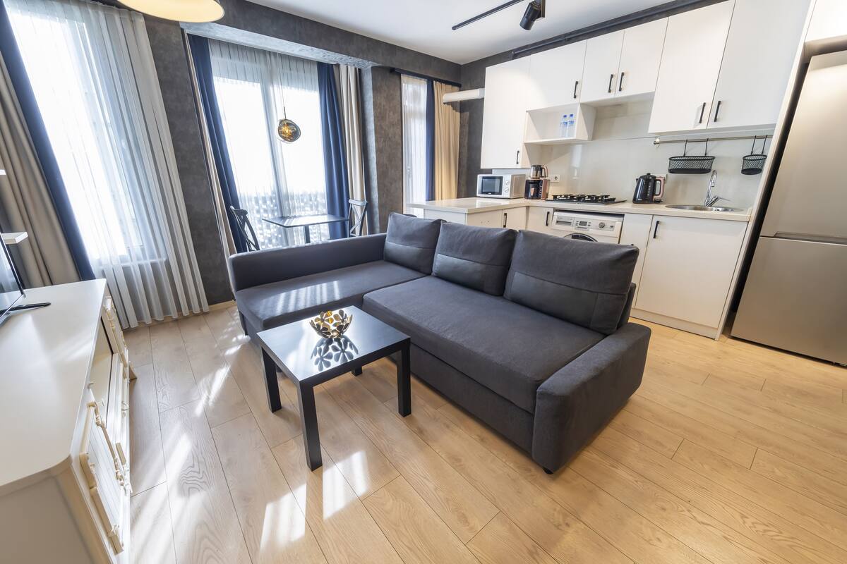 A modern living room with a grey sectional sofa, a small black coffee table, and a kitchen area with white cabinets and stainless steel appliances.