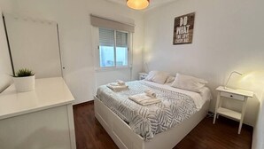 Ein modernes Schlafzimmer mit einem weißen Bett, einer roten Pendelleuchte, einem kleinen Nachttisch und einem Fenster mit Jalousien.