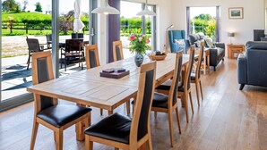 Une salle à manger spacieuse avec une longue table en bois et des chaises, un vase rempli de fleurs et une vue sur un jardin grâce à de grandes fenêtres.