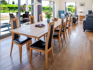 Une salle à manger spacieuse avec une longue table en bois et des chaises, un vase rempli de fleurs et une vue sur un jardin grâce à de grandes fenêtres.