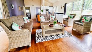A spacious living area with a high ceiling, wicker furniture, and a kitchen in the background. - Natura Turtle Bungalow (Rarotonga)