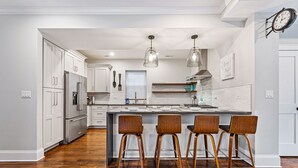 Une cuisine moderne avec des meubles blancs, un îlot central doté de tabourets de bar, des luminaires suspendus et une horloge au mur.
