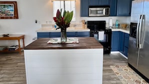 A modern kitchen with blue cabinets, a stainless steel refrigerator, a microwave, and a central island with a vase of flowers. - Afforable & quaint upstairs 2Bedroom 1Bath apartment with lanai and Ocean views (Kailua-Kona)