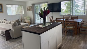 A modern living room with a sofa, a dining table with chairs, a kitchen island, and a TV mounted on the wall. - Afforable & quaint upstairs 2Bedroom 1Bath apartment with lanai and Ocean views (Kailua-Kona)