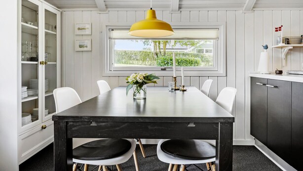 A dining area with a dark wooden table, white chairs, a yellow pendant light, and a window with a view of greenery. - Awesome home in Egernsund with kitchen (Egernsund)