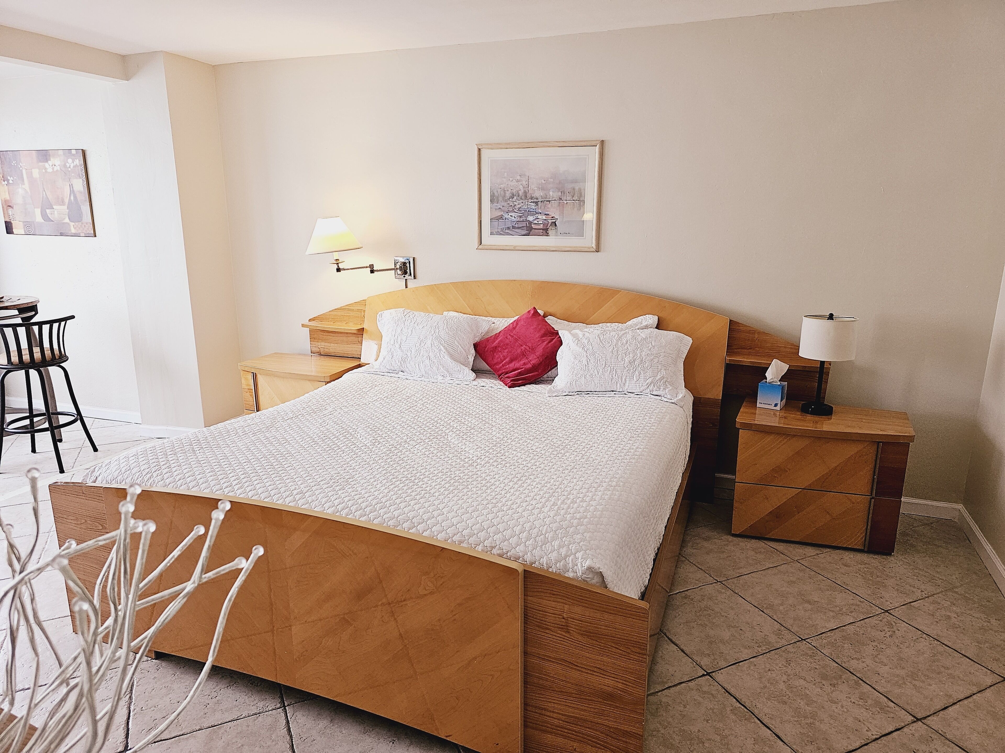 A neatly arranged bedroom with a wooden bed, white bedding, a bedside lamp, and a small table with a tissue box.