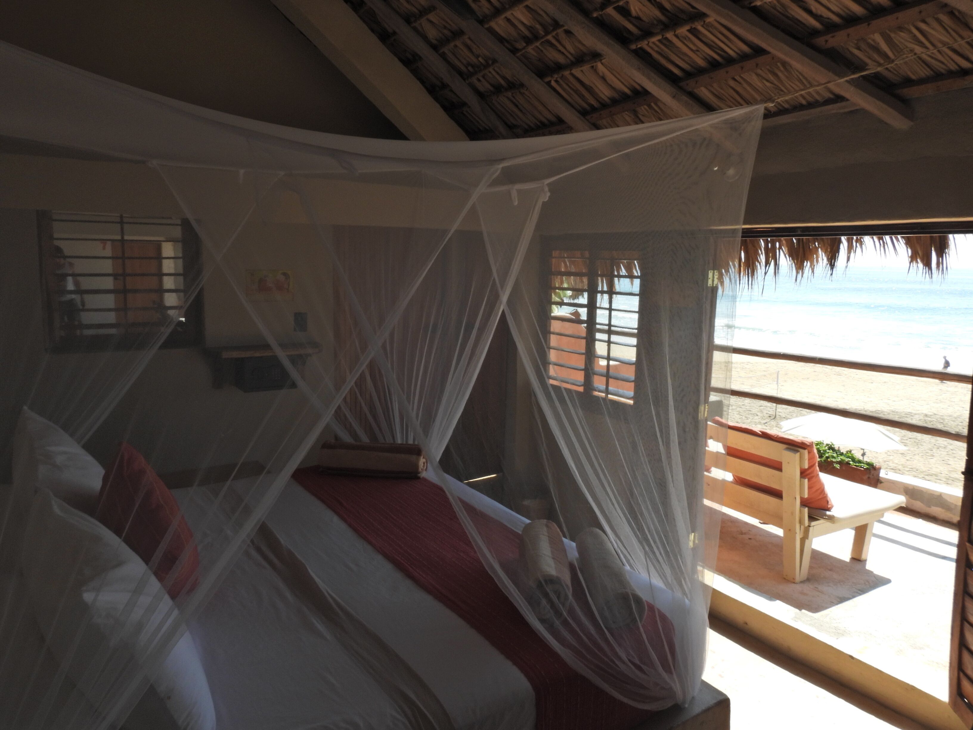 A room with a bed under a mosquito net, a window with a view of the beach, and a wooden bench outside.