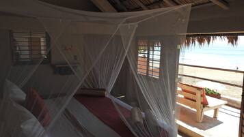 A room with a bed under a mosquito net, a window with a view of the beach, and a wooden bench outside.