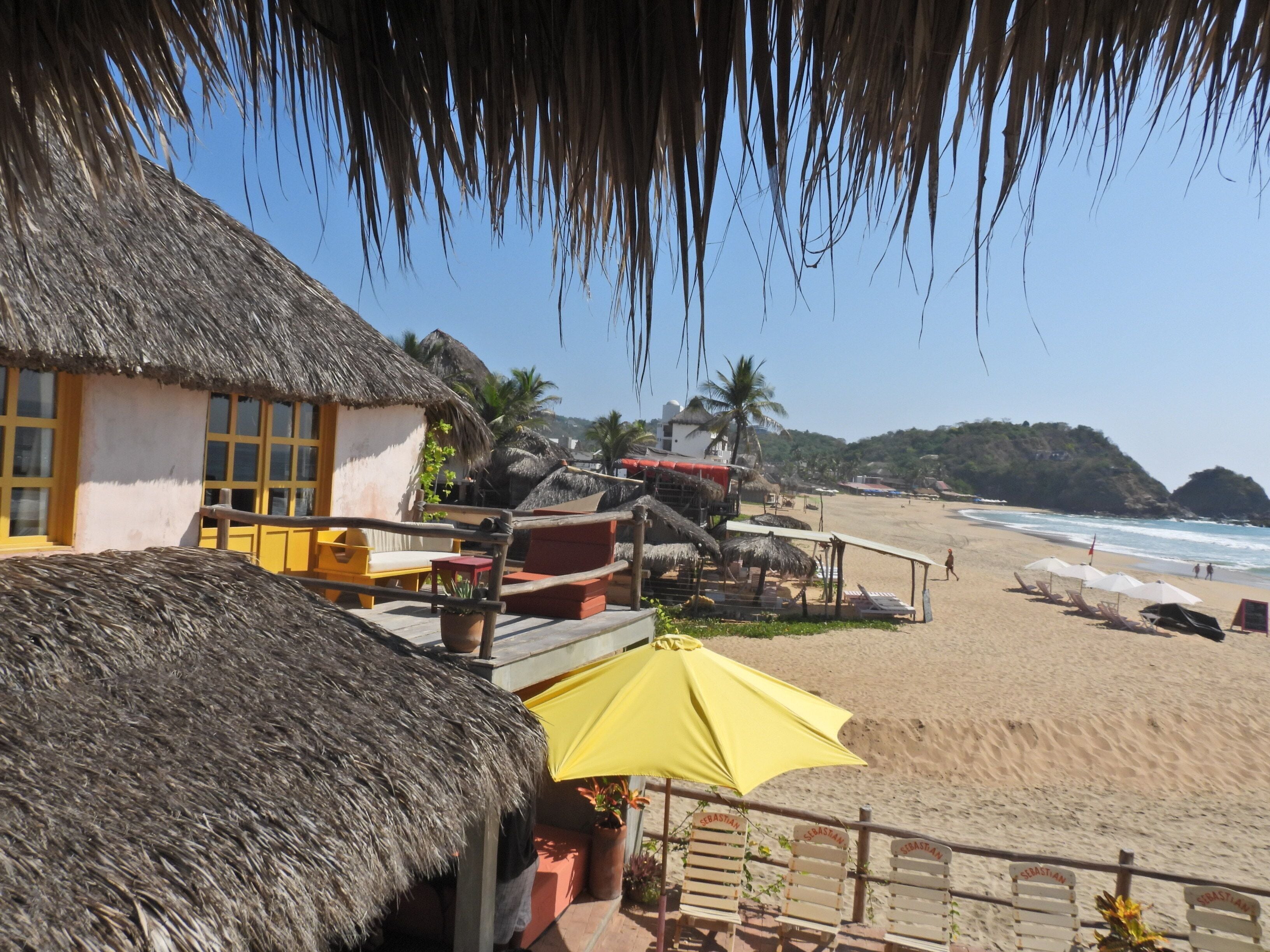 A beachside resort with thatched-roof buildings, sun loungers, and a clear view of the ocean.