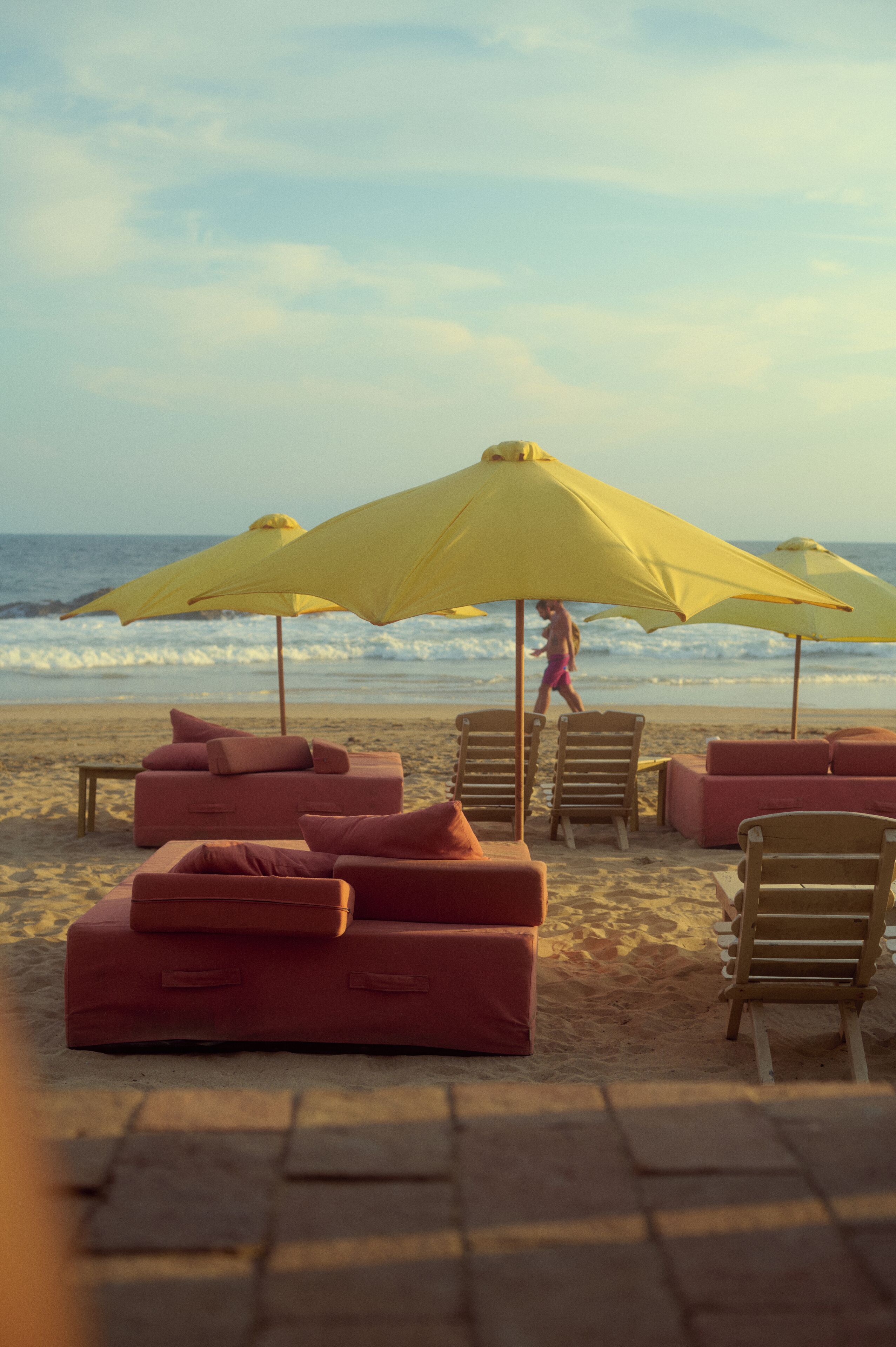 Un área de descanso junto a la playa con varios sombrillas amarillas, asientos acolchados rojos y sillas de madera.