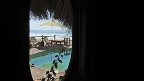 Depuis la fenêtre, on aperçoit un salon de plage avec une piscine, des chaises longues et une table placée sous un toit de chaume.