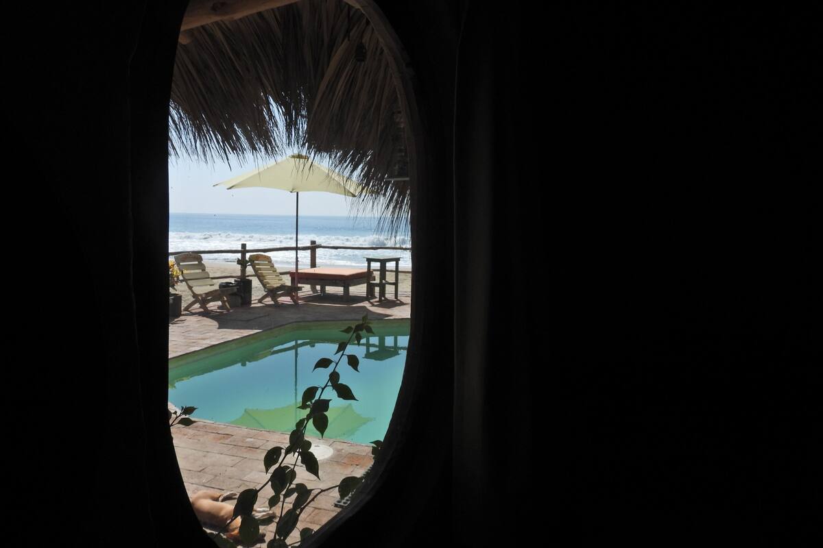 Depuis la fenêtre, on aperçoit un salon de plage avec une piscine, des chaises longues et une table placée sous un toit de chaume.
