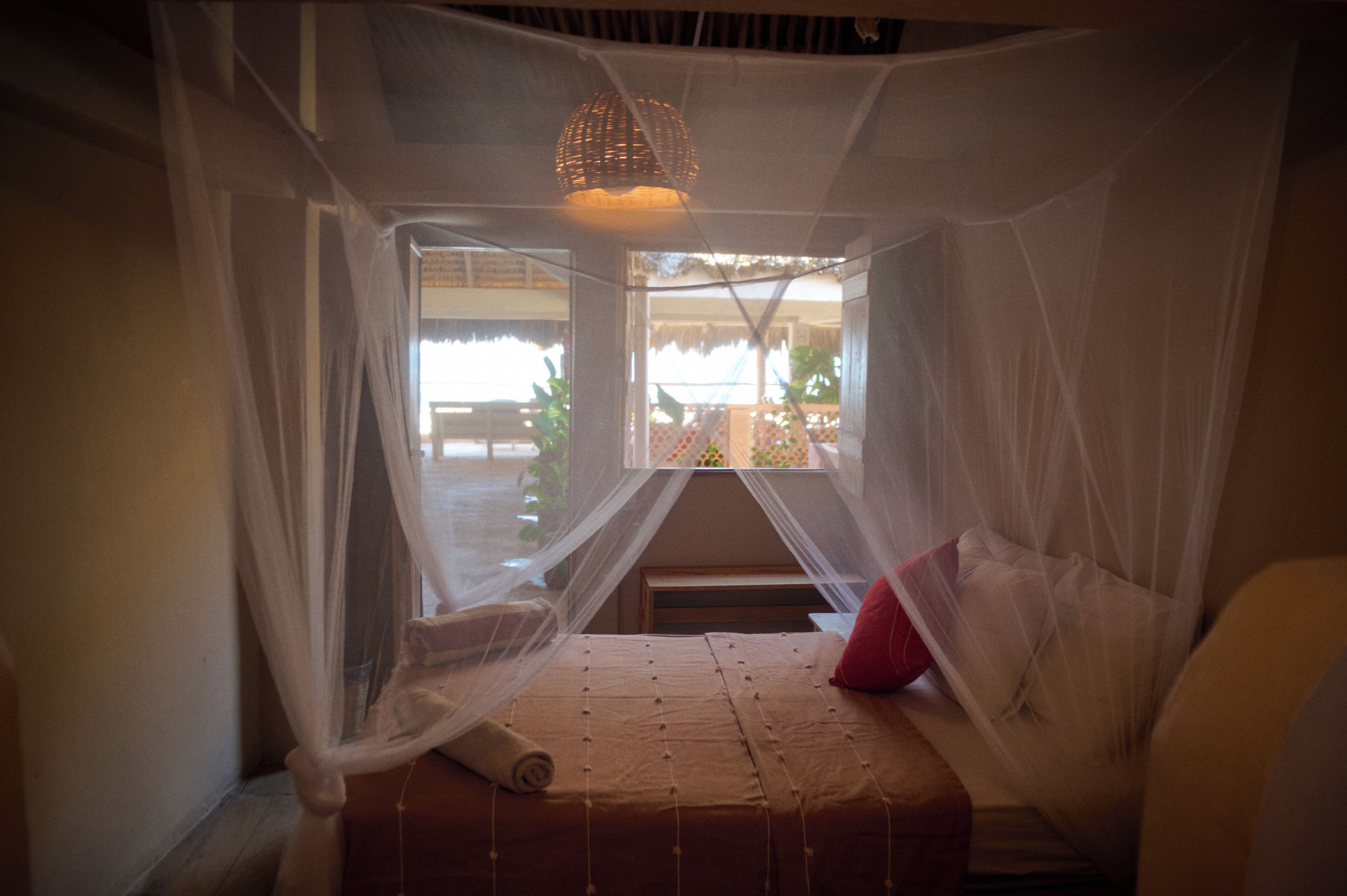 A room with a bed under a mosquito net, a window with a view of the outside, and a hanging light fixture.
