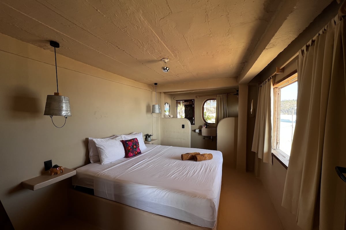 Une chambre avec un grand lit, une table de chevet, un luminaire suspendu, une fenêtre avec des rideaux et une vue sur l’extérieur.