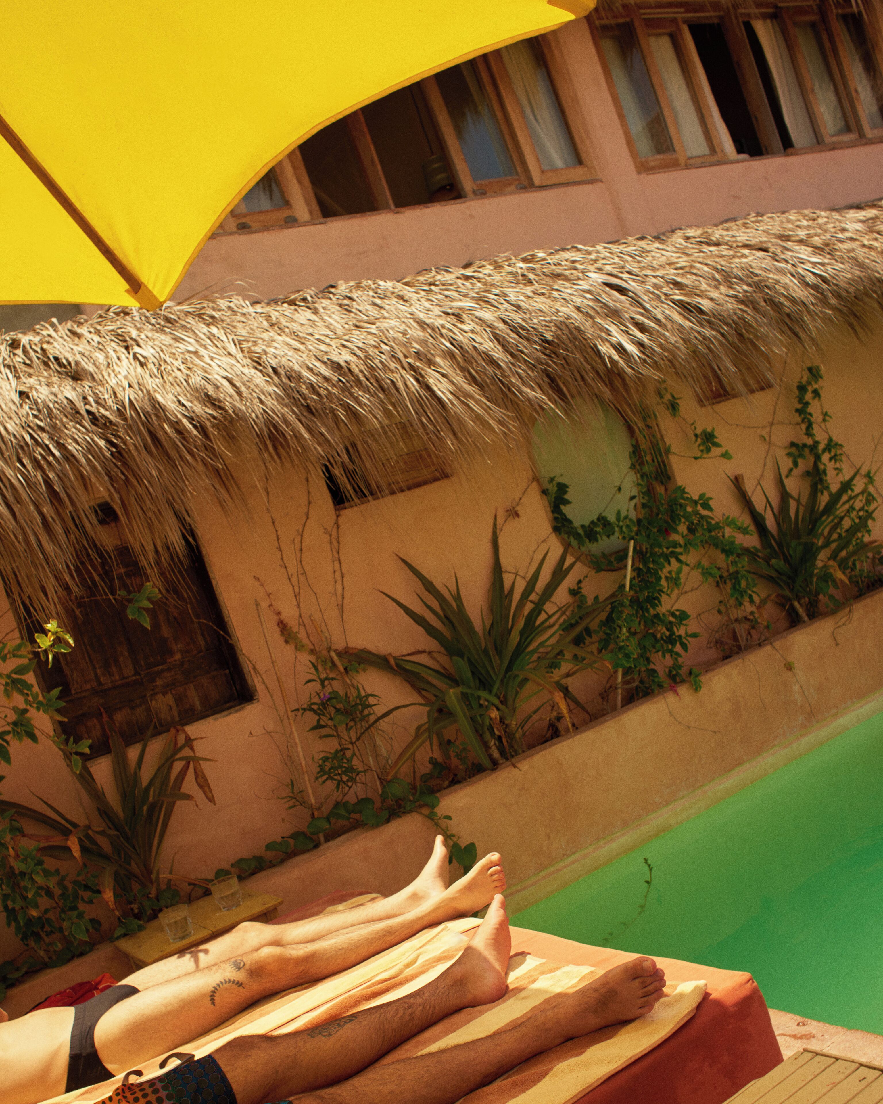 A poolside area with a thatched roof, a wooden bench, and a person relaxing with feet up.