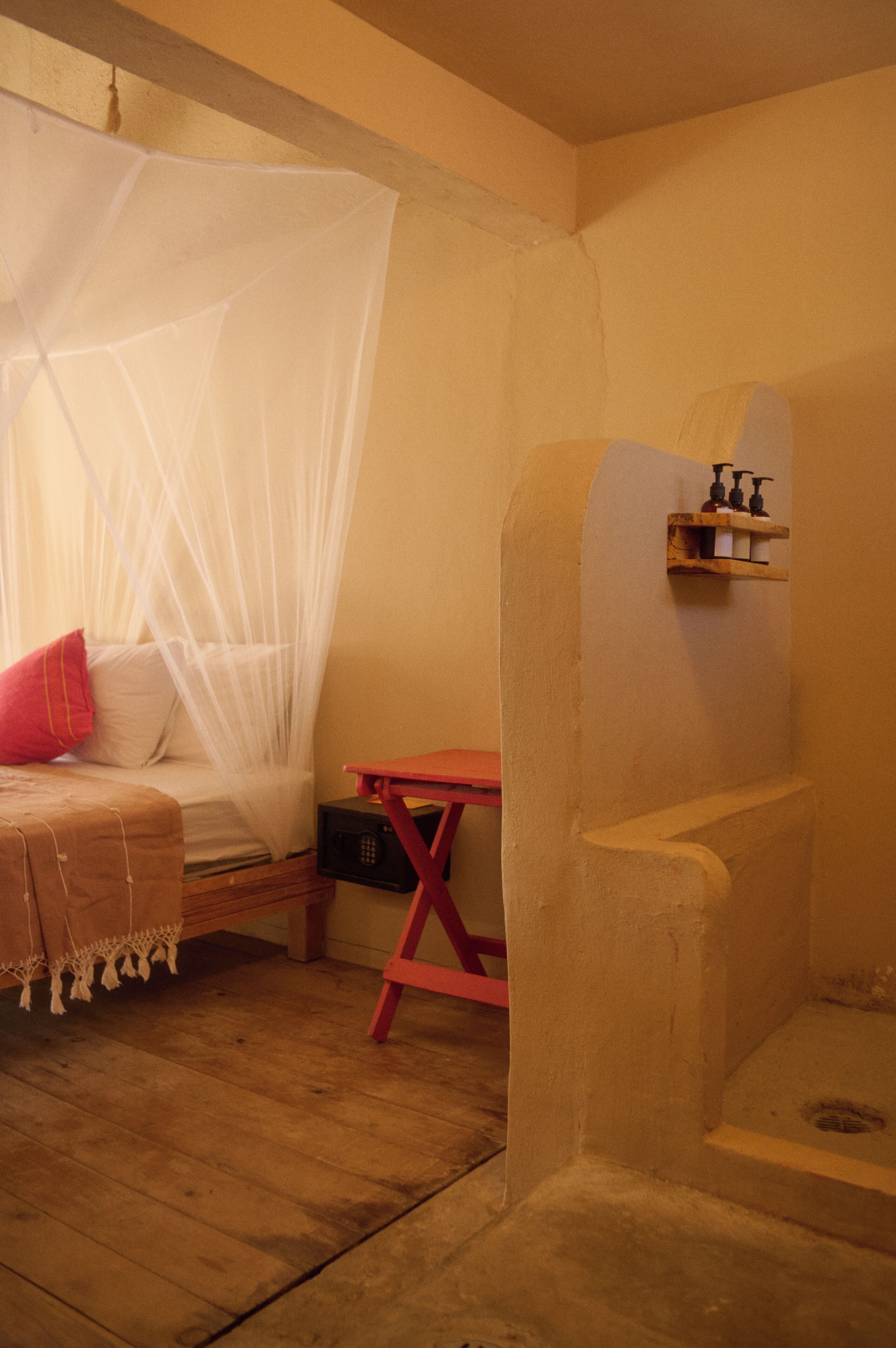 Quarto com cama sob uma rede mosquiteira, piso de madeira, uma cadeira vermelha e uma pequena prateleira com artigos de higiene pessoal.