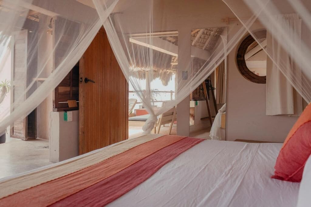 Quarto com cama, porta de madeira, cortina branca e vista para o exterior.