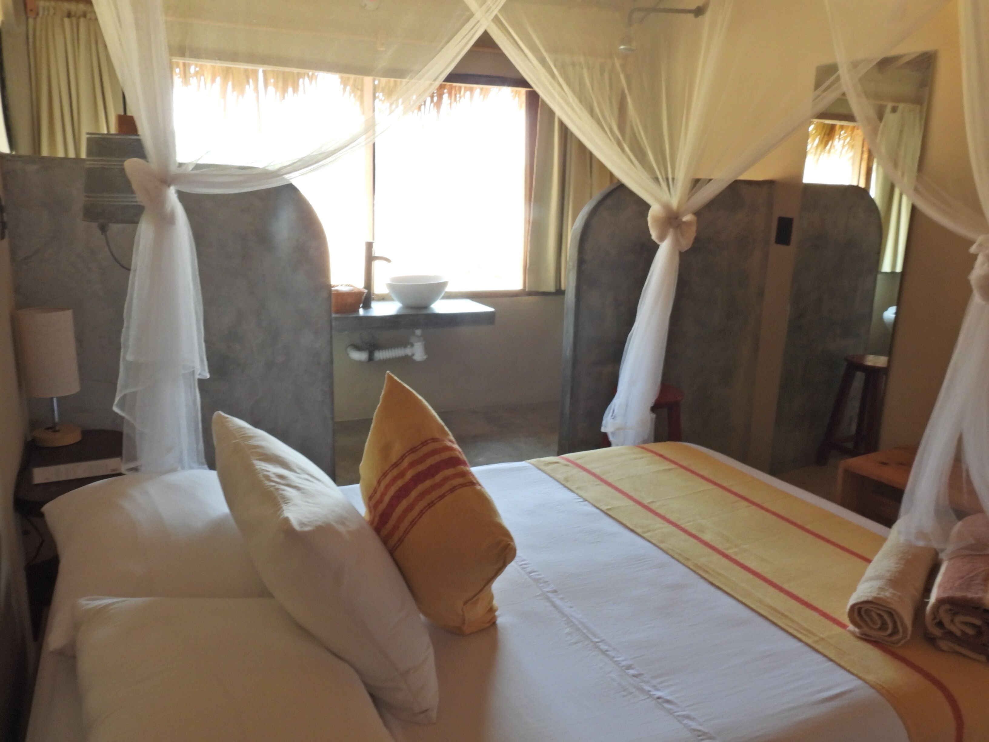 Une chambre d’hôtel avec un lit, une table de chevet, une lampe, une fenêtre avec des rideaux et une salle de bain visible au fond.