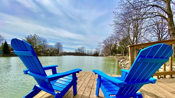 Two blue Adirondack chairs on a wooden deck by a calm lake with trees in the background. - Creekside Cottage - Waterfront Retreat with Kayaks, Fishing & Game Room (Kingsville)