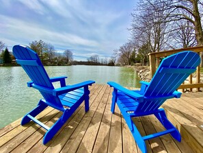 Two blue Adirondack chairs on a wooden deck by a calm lake with trees in the background.