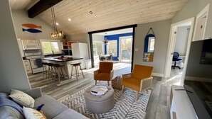 A modern kitchen with white appliances, a dining area with a gray sofa and orange chairs, and a large window providing natural light. - Creekside Cottage - Waterfront Retreat with Kayaks, Fishing & Game Room (Kingsville)