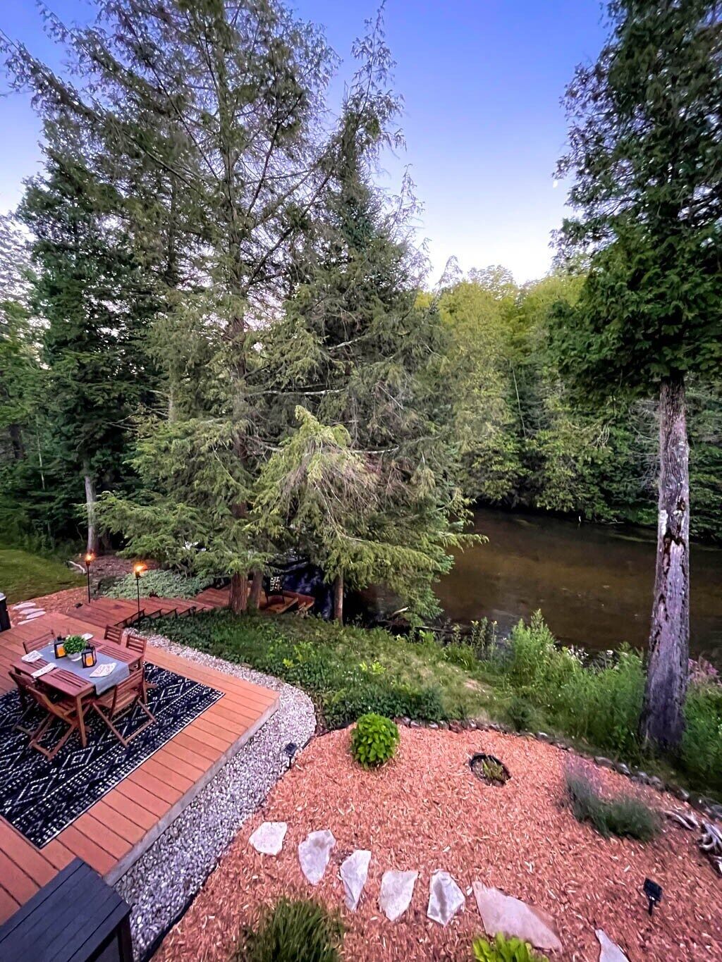 Um deck externo com mesa e cadeiras, cercado por uma floresta e um rio.