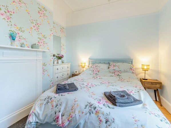 Une chambre à coucher avec une couvre-lit à motifs floraux, une commode avec des tiroirs, un miroir, deux lampes de chevet et une table de chevet avec une lampe.