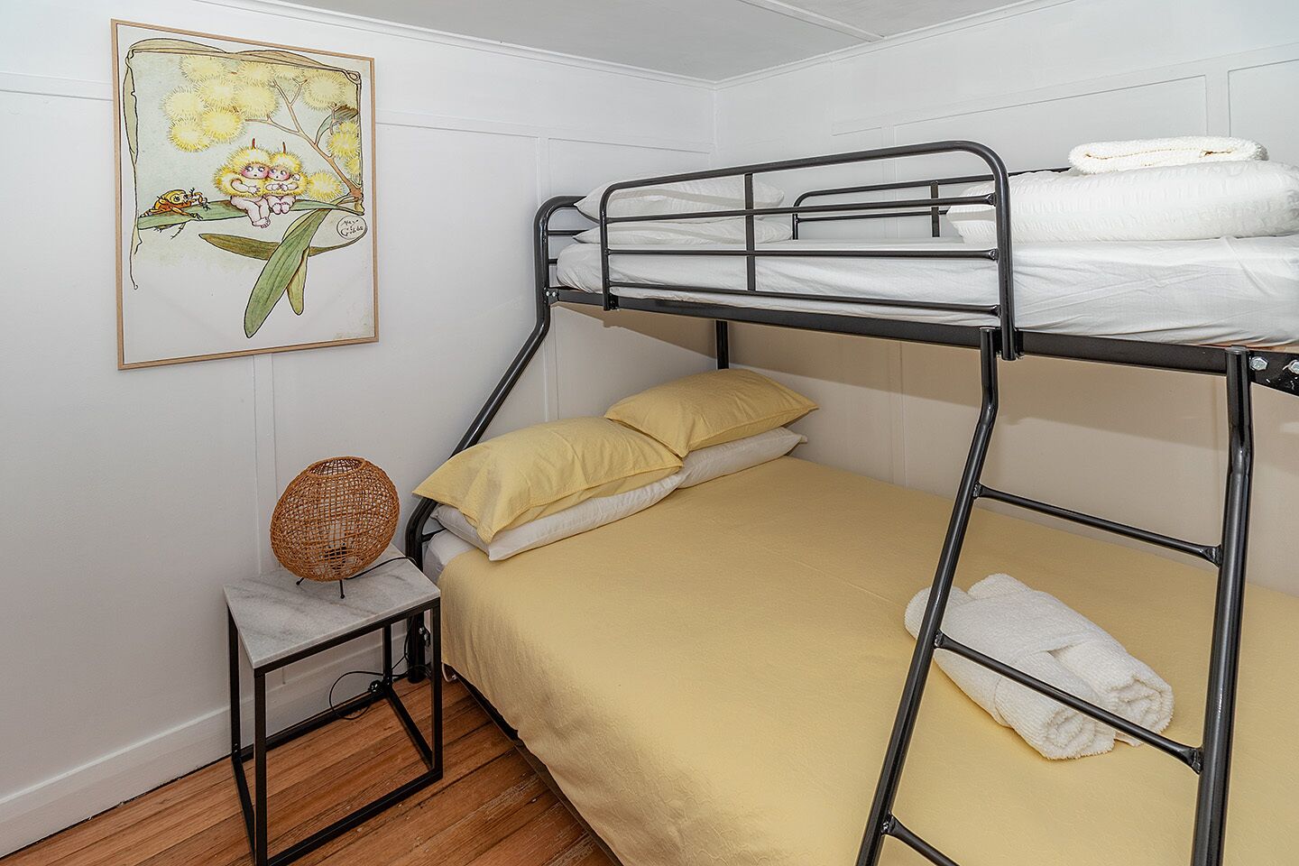 Une chambre avec un lit superposé, une petite table, un panier décoratif et un tableau encadré au mur.