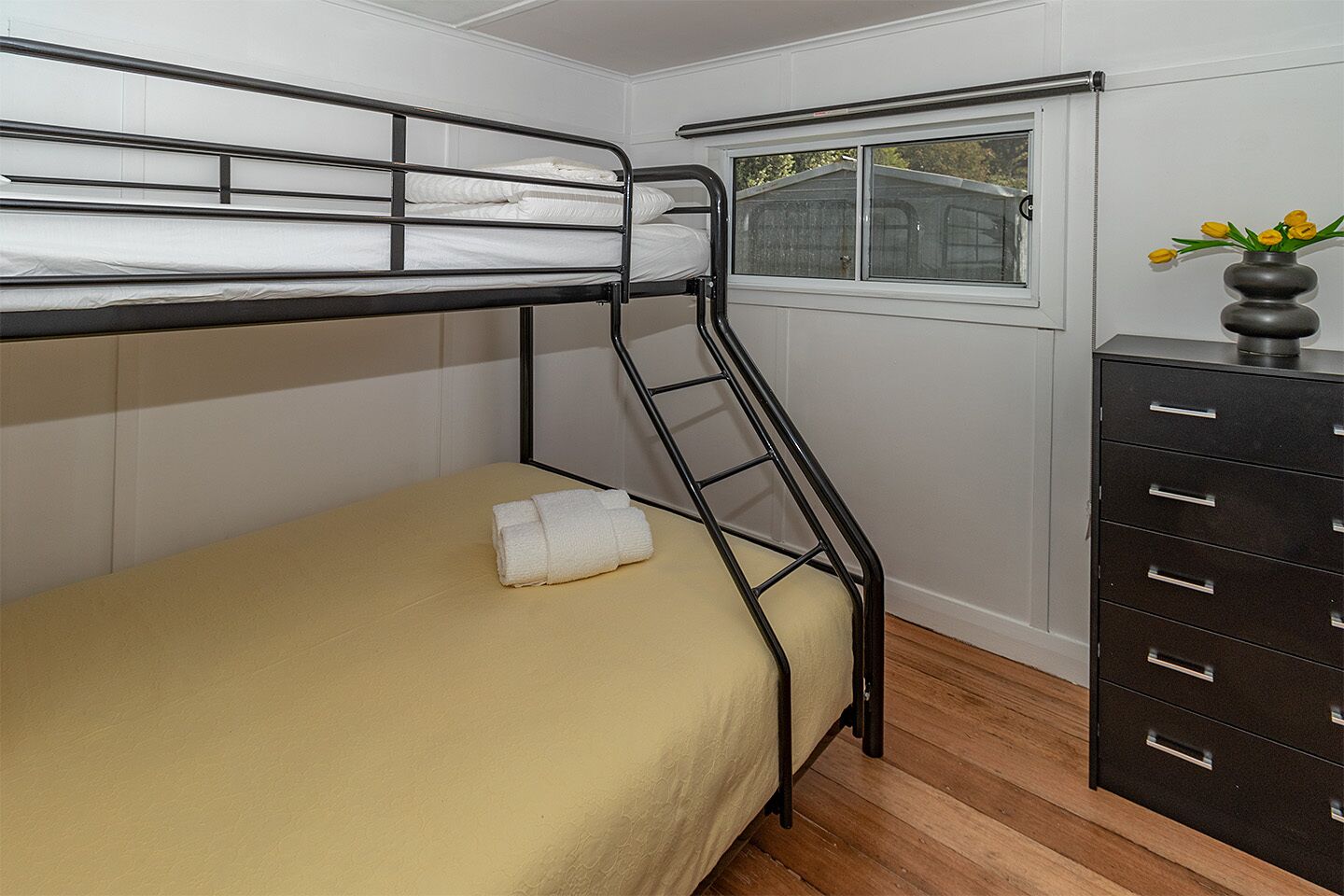 Un lit superposé avec une échelle, une fenêtre donnant sur une maison et une commode avec des tiroirs.