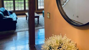 A spacious living room with a large window, a staircase, and a round mirror on the wall. - The Wintergreen Treehouse (Nellysford)