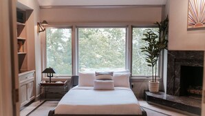 Een slaapkamer met een groot bed, een open haard en uitzicht op bomen door drie ramen.