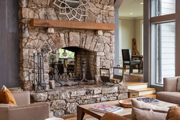 A stone fireplace with a mantel, surrounded by seating and a view of a dining area.
