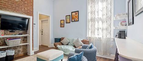 A living room with a brick wall, a flat-screen TV, a ceiling fan, a sofa set, a coffee table, and a window with sheer curtains.