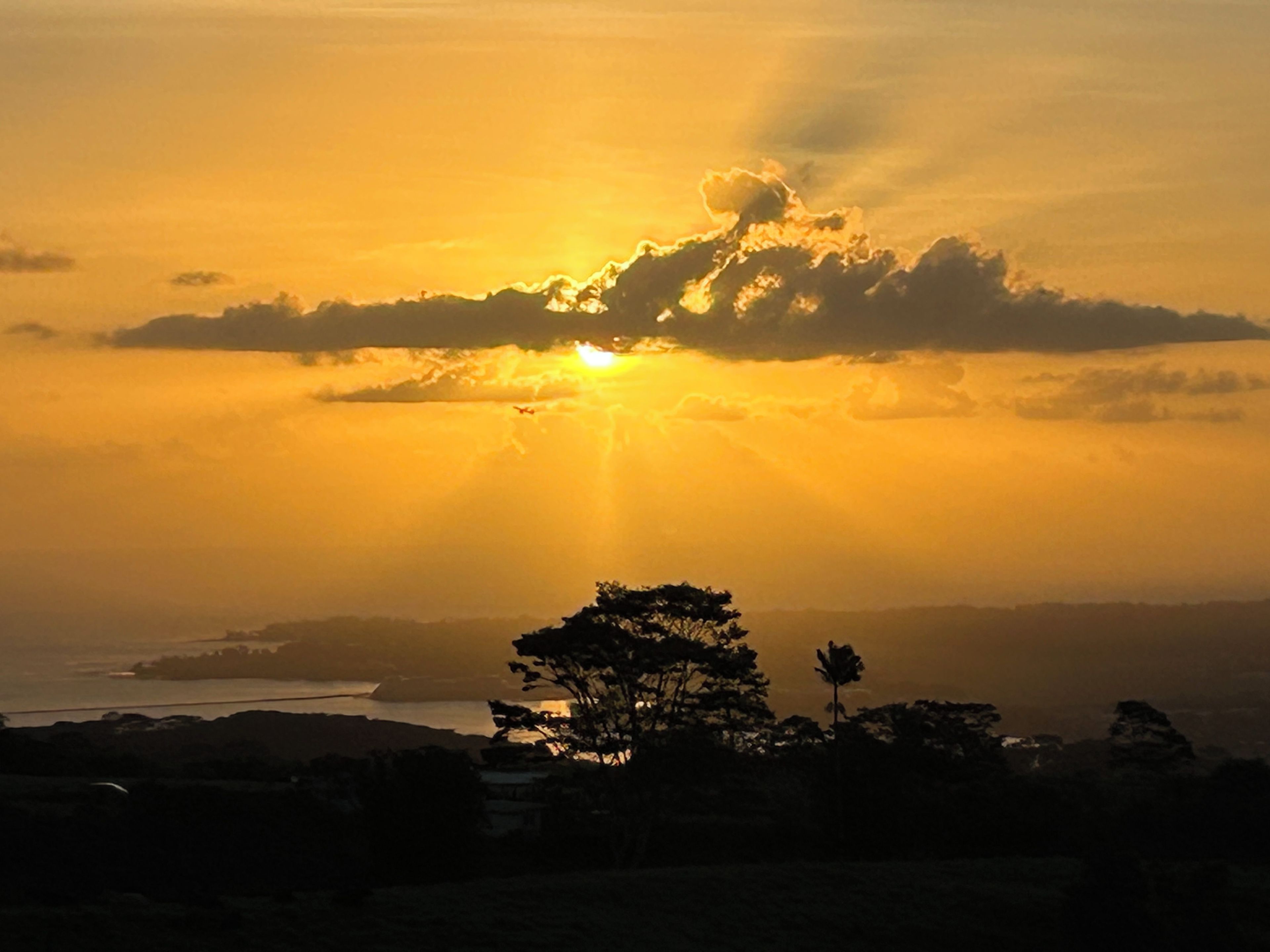 A sunset over a coastal landscape with silhouettes of trees and distant landforms.